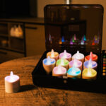 Zestaw 12 podgrzewaczy LED RGB DORSETTO w otwartym czarnym etui na okrągłym drewnianym stole – jeden podgrzewacz ustawiony poza etui, w tle nowoczesny salon